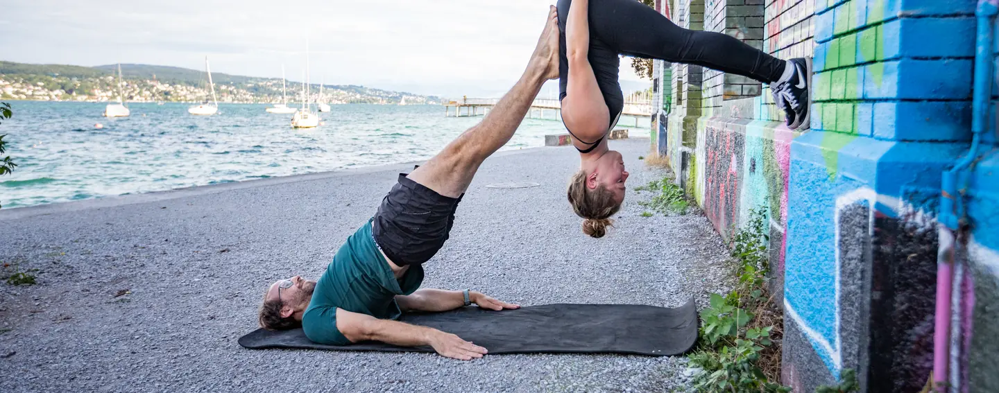 A base lying on a mat on the shore of Lake Zürich holding a flyer upside down on his feet, sailboats moored behind them.