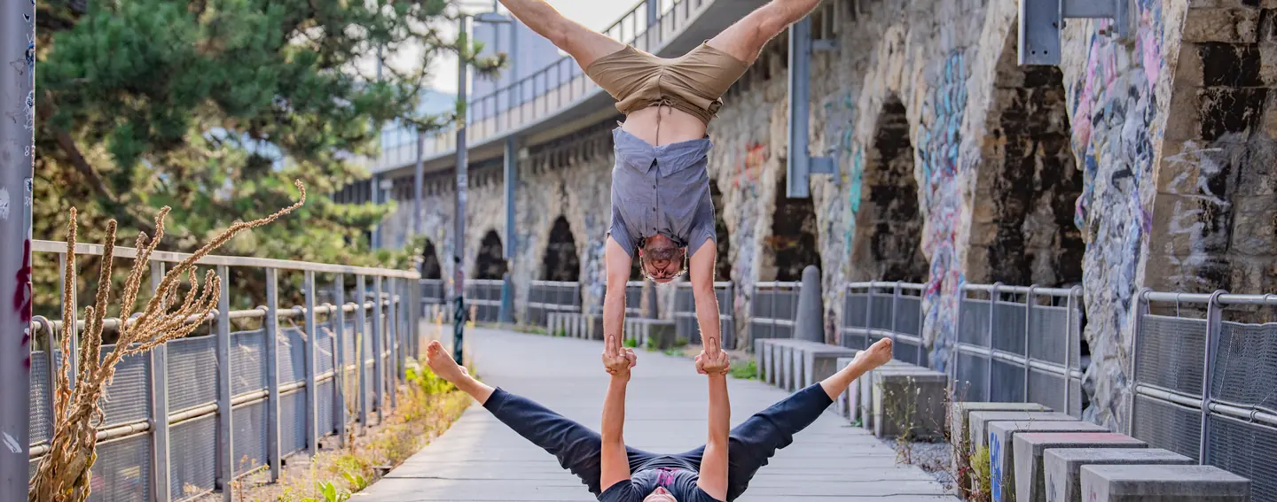 A flyer in a straddle handstand on her base's hands, on the riverside promenade at the Letten in Zürich.